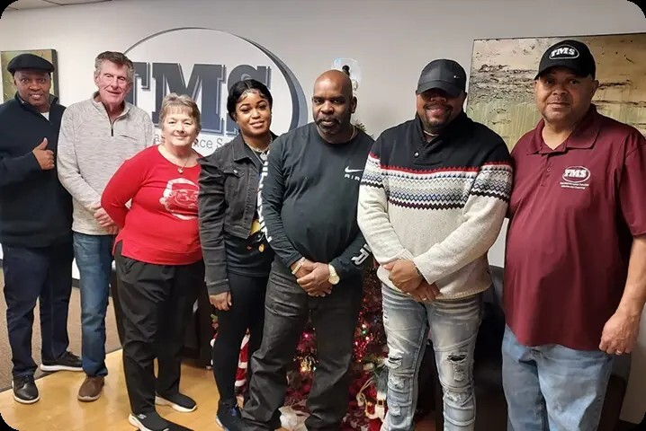 A group of 7 people smiling and standing in front of the TMS logo